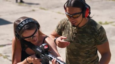 Bearded man in camo t-shirt watching woman aiming submachine gun. Safety headphones and goggles. Firearms training at firing range. Outdoor horizontal shot. High quality 4k footage