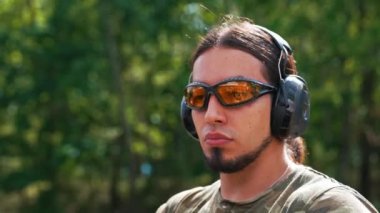 Man wearing camouflage t-shirt safety headphones and goggles aiming machine gun. Outdoor shooting range target practise. Horizontal shot. High quality 4k footage