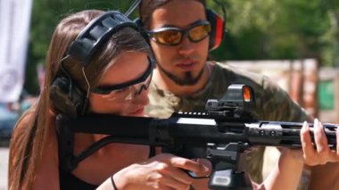 Caucasian woman in safety headphones and goggles aiming submachine gun watched by bearded man in camouflage t-shirt. Firearms training ar fiiring range. Horizontal shot. High quality 4k footage