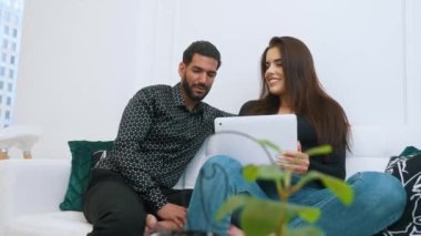 Man with dark hair and beard smiling sitting with his wife on couch browsing internet on laptop. Young couple conversation. Indoor High quality 4k footage