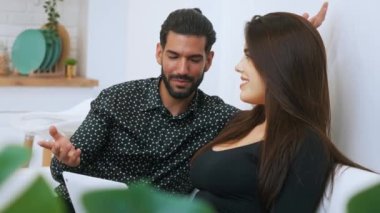 White woman with long dark hair working on laptop listening to her partner talking to her and smiling sitting on couch together. Expressive conversation. Young couple. Indoor. High quality 4k footage