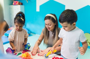 Relaxing sensory play with moldable kinetic sand at nursery school. Toddlers with their teacher having fun around the table using different tools for sculpting sand such as colorful and textured