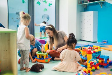 Toddlers and their nursery teacher playing with plastic building blocks and colorful car toys while sitting on the floor in a playroom. Concentration, fine motor and gross motor skills development