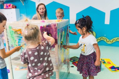 Cling wrap painting for improving kids imagination and brain development. Toddlers and their teacher painting with brushes, rollers and paints on a cling film wrapped around the wooden shelving stand