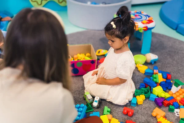Learning through play at the nursery school. Toddler little girl and the teacher playing with colorful plastic playhouses, building blocks, cars and boats. Imagination, creativity, fine motor and
