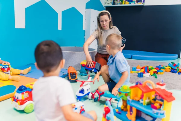 Toddlers and their nursery teacher playing with plastic building blocks and colorful car toys in a nursery school playroom. Early brain and motor skills development. High quality photo