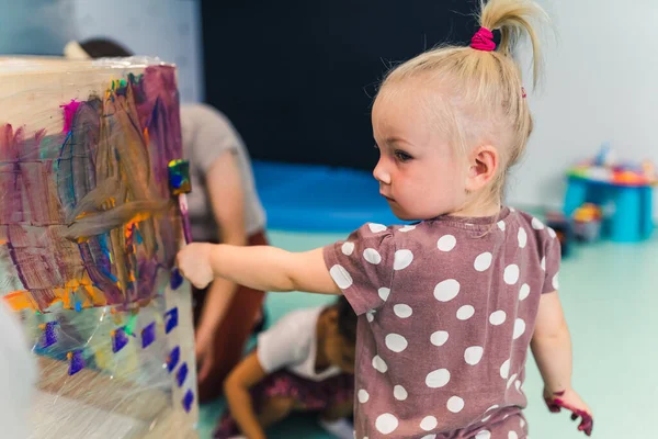 Cling film painting. Little caucasian girl toddler painting with her arms on a cling film wrapped all the way round the wooden shelf unit. Creative activity for kids development at the nursery school
