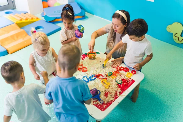 Fun sensory play with moldable kinetic sand at nursery school. Toddlers standing around the table and using different tools for sculpting sand such as colorful and textured rolling pins, cutters
