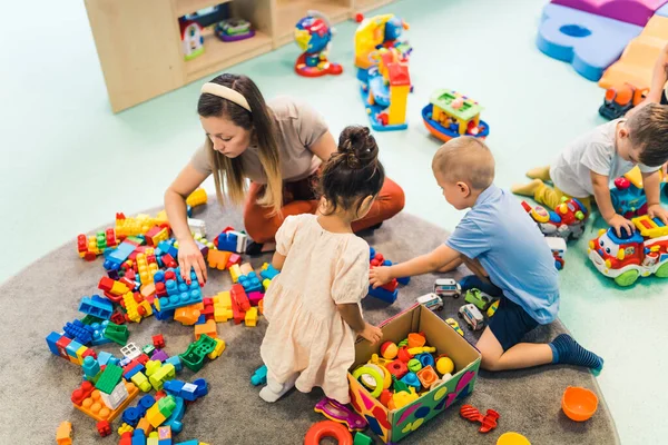 Toddlers and their nursery teacher playing with plastic building blocks and colorful car toys while sitting on the floor in a playroom. Concentration, fine motor and gross motor skills development