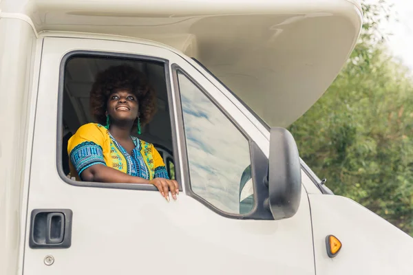 Dark-skinned smiling African young woman with afro haircut, wearing a bright boho outfit, looking out of the window of a camper van and admiring peaceful nature around at the end of her road trip