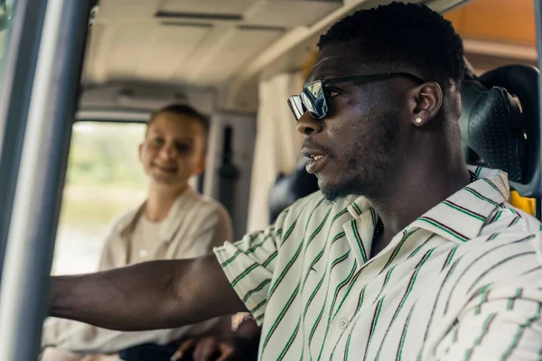 Dark-skinned young man driver close up. Multi-ethnic group of close friends having a summer road trip in a camping van, driving, talking and laughing heartily inside their vehicle. Front seats close