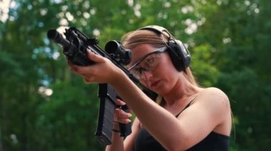 Young caucasian woman wearing protective goggles and headphones aiming submachine gun on training ground. Blurred background horizontal shot. High quality 4k footage