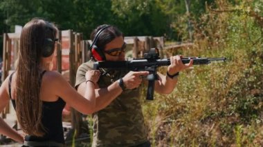 Woman standing next to man aiming submachine gun both wearing safety headphones and goggles. Firearms training at firing range. Outdoor horizontal shot. High quality 4k footage