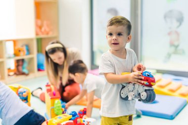 Toddlers and their nursery teacher playing with plastic building blocks and colorful car toys while sitting on the floor in a playroom. Concentration, fine motor and gross motor skills development