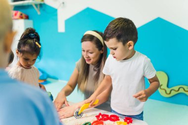 Calming sensory play with moldable kinetic sand at nursery school. Toddlers with their teacher having fun around the table using different tools for sculpting sand such as colorful and textured