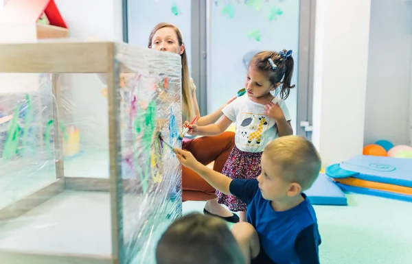 Cling wrap painting for improving kids imagination and brain development. Toddlers and their teacher painting with brushes, rollers and paints on a cling film wrapped around the wooden shelving stand