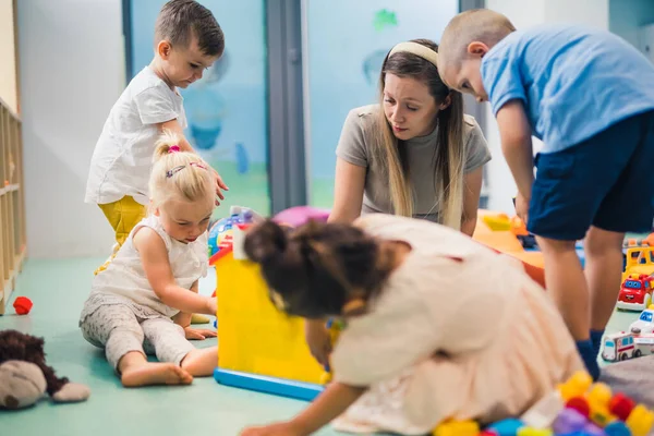 Toddlers and their nursery teacher playing with plastic building blocks and colorful car toys while sitting on the floor in a playroom. Concentration, fine motor and gross motor skills development