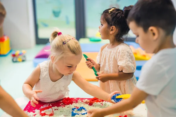 Fun sensory play with moldable kinetic sand at nursery school. Toddlers standing around the table and using different tools for sculpting sand such as colorful and textured rolling pins, cutters