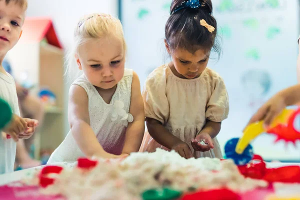 Relaxing sensory play with moldable kinetic sand at nursery school. Toddlers having fun around the table using different tools for sculpting sand such as colorful and textured rolling pins, cutters