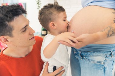 Little caucasian child son supported by his young adult father giving a kiss to his mothers pregnant belly. Siblings concept. High quality photo