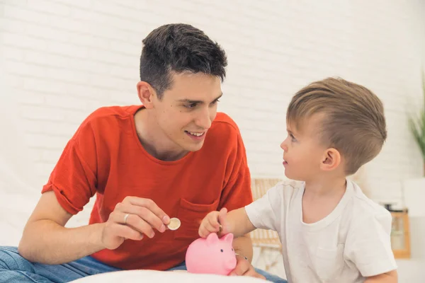 Savings concept. Proud young adult caucasian dark-haired father in orange t-shirt holding a coin and teaching his young preschooler son how to use piggybank. High quality photo