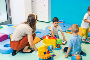 Toddlers and their nursery teacher playing with plastic building blocks and colorful car toys while sitting on the floor in a playroom. Concentration, fine motor and gross motor skills development