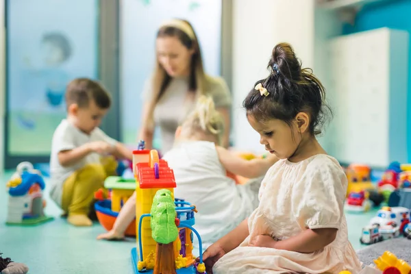 Learning through play at the nursery school. Toddler little girl and the teacher playing with colorful plastic playhouses, building blocks, cars and boats. Imagination, creativity, fine motor and