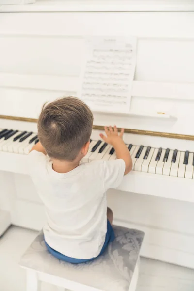 Music in the kids development. Vertical indoor shot of a young caucasian boy in white t-shirt sit-in by the piano and trying to play some music. High quality photo