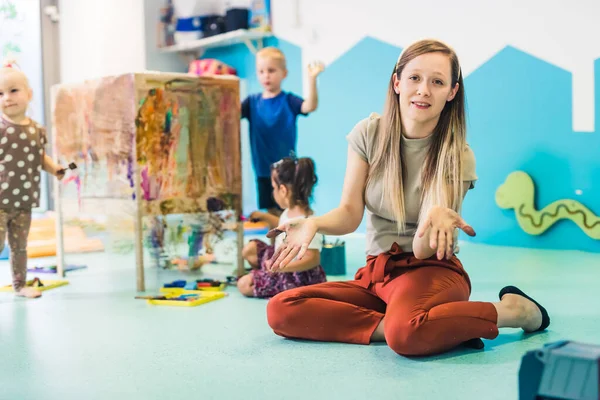 Young caucasian day-care-center teacher sitting on the floor and showing her arms to the camera while on the background toddlers painting with tempera paints on a cling film wrapped around the wooden