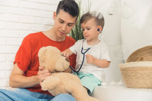 Different games with children. Medium closeup indoor shot of caucasian millennial dad with his preschooler son playing doctor with teddy bear. Stethoscope. High quality photo
