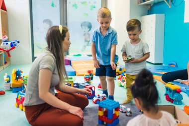Toddlers and their nursery teacher playing with plastic building blocks and colorful car toys in a nursery school playroom. Early brain and skills development. High quality photo