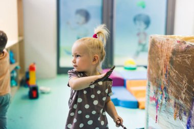 Cling film painting. Little caucasian girl toddler painting with her arms on a cling film wrapped all the way round the wooden shelf unit. Creative activity for kids development at the nursery school