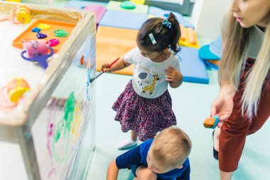 Cling film painting for improving kids imagination. Toddlers painting with, brushes, rollers and paints on a cling film wrapped around the wooden shelving stand. Fun activity for kids sensory skills