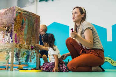 Cling film painting. Toddlers painting with a sponge, brushes and paints on a cling film wrapped all the way round the wooden shelf unit. A teacher helping them. Creative activity for kids sensory