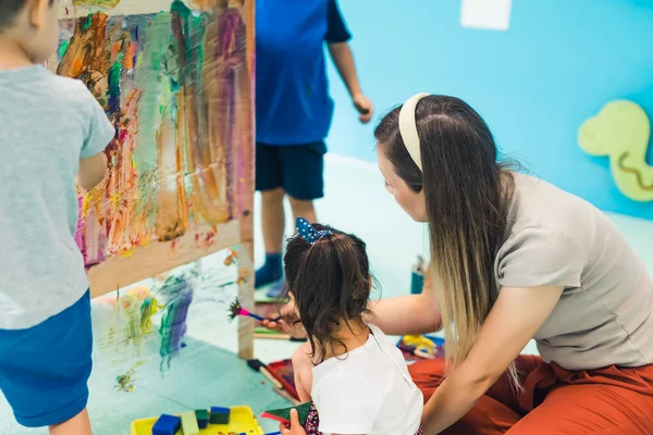 Cling film painting. Toddlers painting with a sponge, brushes and paints on a cling film wrapped all the way round the wooden shelf unit. A teacher helping them. Creative activity for kids sensory