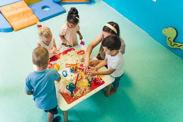 Calming sensory play with moldable kinetic sand at nursery school. Toddlers with their teacher having fun around the table using different tools for sculpting sand such as colorful and textured
