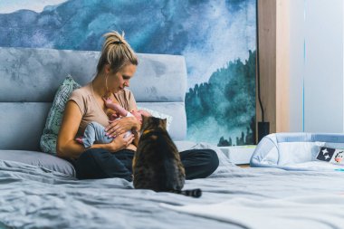 Bekar bir anne, bebeğini beslemek için yatakta bacak bacak üstüne atarak oturuyor. Tekir kedi ilginç bir şekilde yeni bir aile üyesine bakıyor. Yatak odasının içi. Yüksek kalite fotoğraf