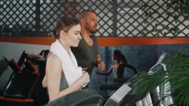 Boynunda havluyla beyaz kadın ve spor salonunda koşu bandında koşan tişörtlü kaslı adam. Kardiyo egzersizi. Yatay video. Yüksek kalite 4k görüntü