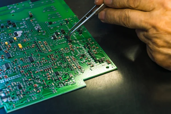 Hand gripping tweezers assembling electrical parts on detailed surface of printed circuit board PCB. Repairing technology. Precise. Close-up horizontal shot. High quality photo
