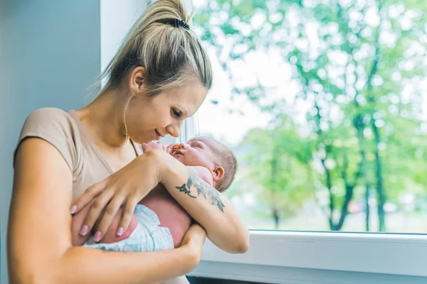 Lovely mother and infant relationship. Calm caucasian tattooed blonde girl in beige t-shirt holding her little baby boy in her arms while standing next to the window. High quality photo
