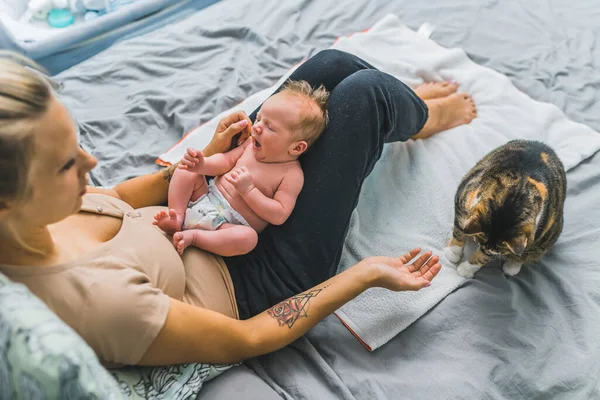 Middle-aged caucasian tattooed mother holding her beautiful yawning infant baby boy in diaper and stretching her arm to pet her tabby cat. New family member. High quality photo