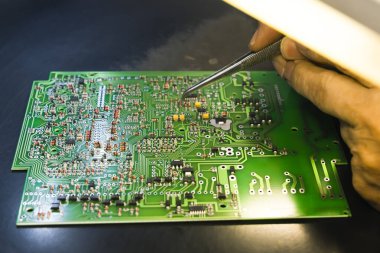 PCB, elektrik bileşenleriyle basılmış devre kartı ve lambayla daha fazla parça yakan cımbızla tutturulmuş bir el. Elektronikleri monte ediyorum. Tamirci. Yatay çekim. Yüksek kalite fotoğraf