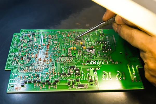 PCB, elektrik bileşenleriyle basılmış devre kartı ve lambayla daha fazla parça yakan cımbızla tutturulmuş bir el. Elektronikleri monte ediyorum. Tamirci. Yatay çekim. Yüksek kalite fotoğraf