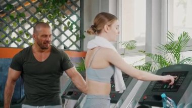 Kafkas saçlı, boynuna havlu bağlanmış beyaz kadın arka planda dinlenen kaslı erkek arkadaşıyla koşu bandını durdurdu. Yatay video. Yüksek kalite 4k görüntü