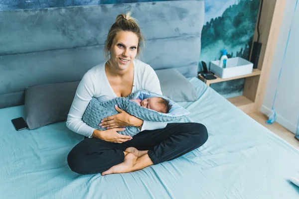 White woman with blonde hair sitting in bed holding her newborn baby wrapped in blanket looking into camera smiling. Proud young mom. Horizontal indoor shot. High quality photo