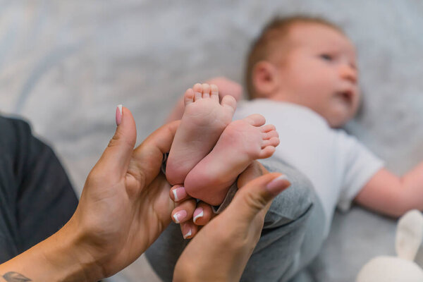 A loving new mother gently holding her infant sons little feet. A baby boy with cute brown hair, laying on his back on a grey blanket with his arms stretched to the sides. High quality photo