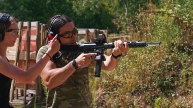 Woman standing next to man aiming submachine gun both wearing safety headphones and goggles. Firearms training at firing range. Outdoor horizontal shot. High quality 4k footage