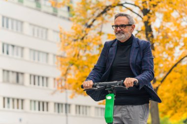 Güz aylarında ilk kez en sevdiği parkta elektrikli scooter kullanan beyaz saçlı, yakışıklı bir adam. Bulanık bina ve arka planda sarı ağaç. Yüksek kalite