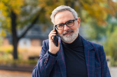 Yakışıklı beyaz saçlı şirket sahibi, kulağının yanında siyah bir akıllı telefon tutuyor ve bir telefon bekliyor. Arka planda bulanık park ağaçları var. Ceket ve balıkçı yaka. Yüksek kalite fotoğraf
