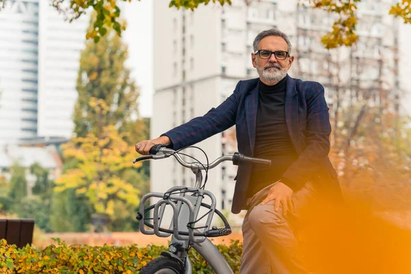 Autumn in the city. Medium long shot of middle-aged bearded man sitting on rented city bike dressed in business casual way. Blurred orange leaf in the foreground. High buildings and colourful leaves